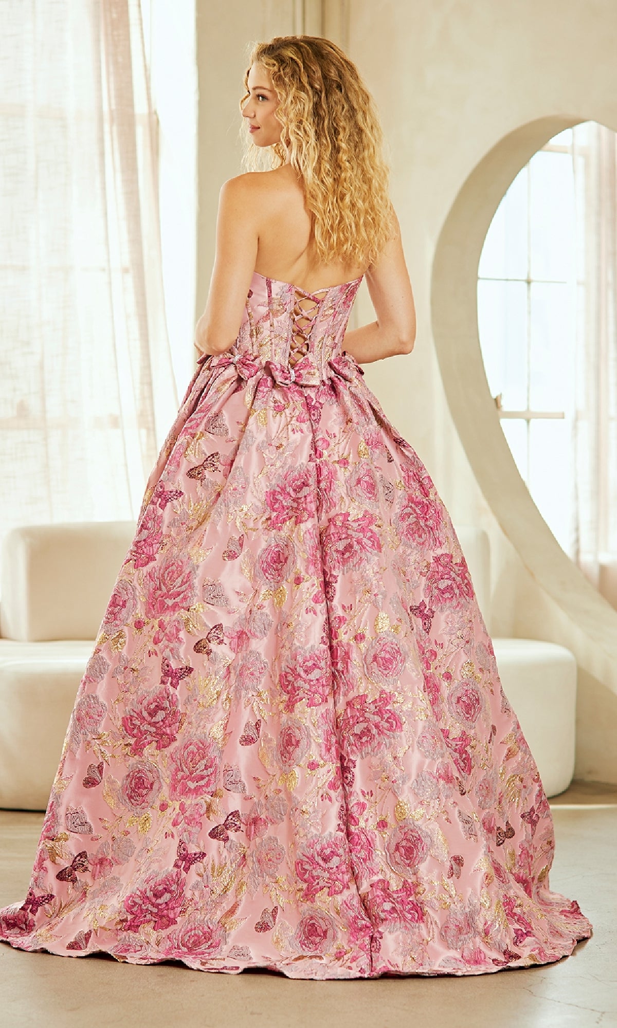 A woman with curly blonde hair stands in a sunlit room wearing the Lace-Up Print Prom Ball Gown: Amelia Couture 8068, a strapless pink floral ball gown featuring a lace-up back and bow.